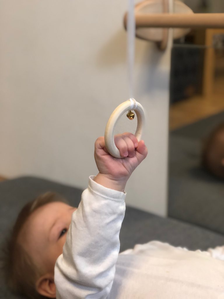 A baby boy has managed to grab and pull the Ring on a Ribbon Tactile Mobile which hangs on an elastic ribbon, Essential Montessori Toys for Babies