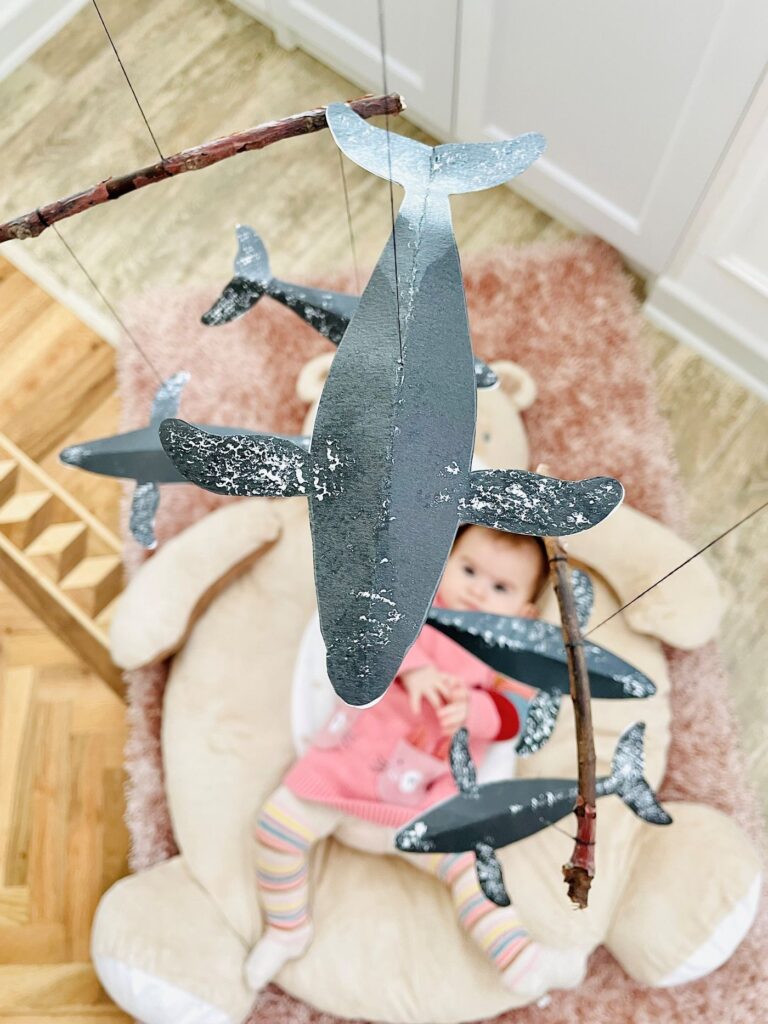 Montessori-inspired black and white whale baby mobile with simple whale silhouettes, watched by a baby girl practicing visual focus and concentration., Bebé observando figuras que se mueven lentamente, desarrollando el seguimiento visual y la concentración temprana.